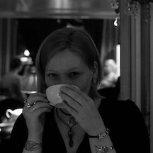 Foto von Klaudia in einem Kaffeehaus, eine weiße Tasse vor dem Gesicht
